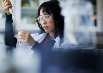 A woman wearing safety goggles and a lab coat uses a pipette to transfer liquid to a test tube in a lab.