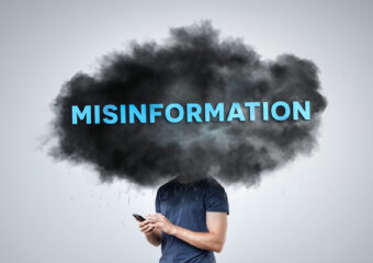 A person stands holding a smartphone, with their head obscured by a dark cloud labeled "Misinformation." The image symbolizes the concept of fake news and digital deception, illustrating the impact of misinformation in the digital age.